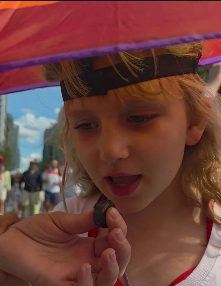 A young girl shelters under a rainbow umbrella at a street gathering, looking down at a pin held in an adult's hand.